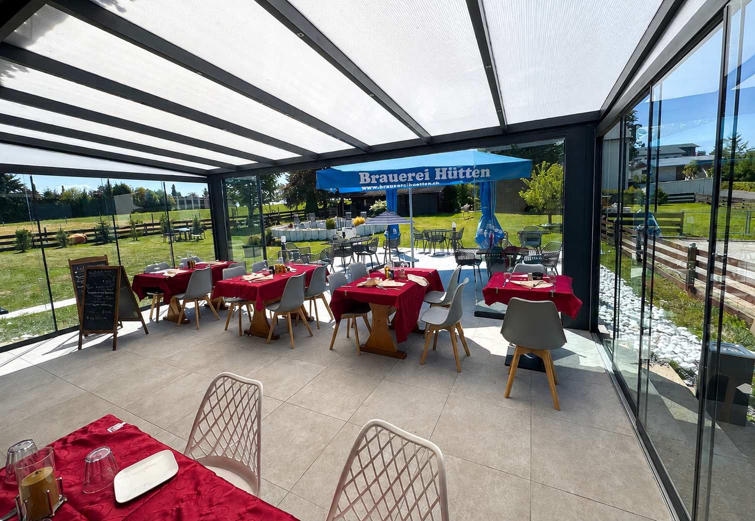 Der verglaste Wintergarten des Bistro Johannes mit eingedeckten Tischen, Stühlen und Blick auf Terrasse und Garten